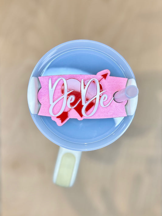 Close-up of a pink Valentine’s Day tumbler topper with a red heart and arrow, personalized with the name “DeDe” in white script, on a white/clear tumbler lid